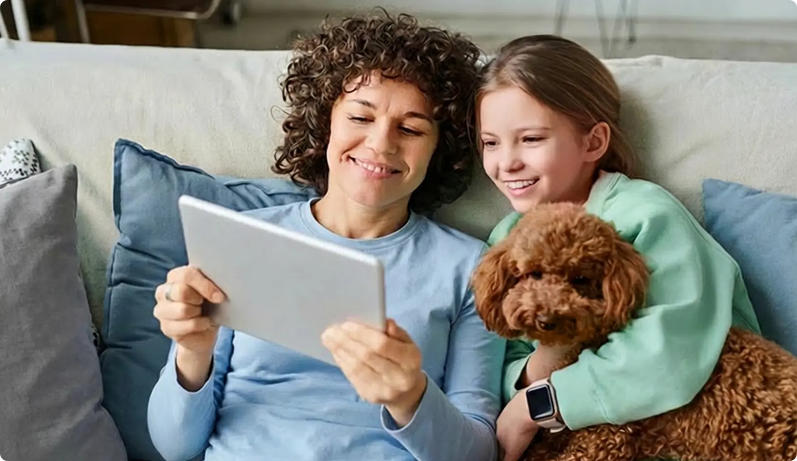 Žena a dievča sedia na gauči doma, usmievajú sa a pozerajú sa na tablet, pričom dievča drží malého psa.