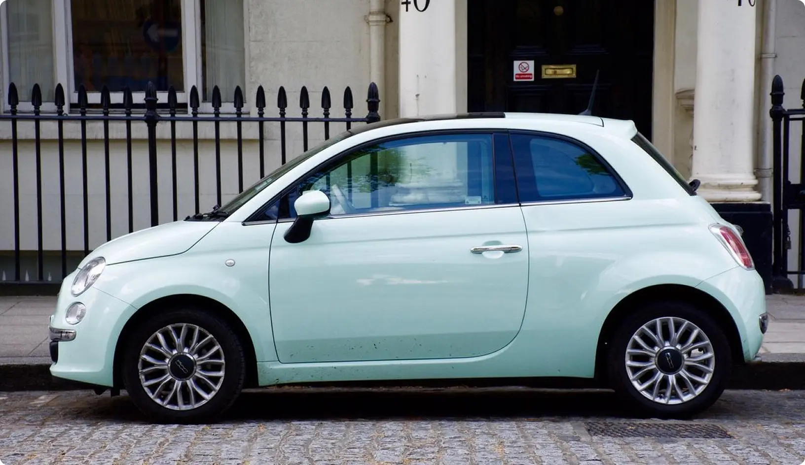 Svetlomodré malé mestské auto zaparkované pri chodníku pred budovou s čiernym plotom.