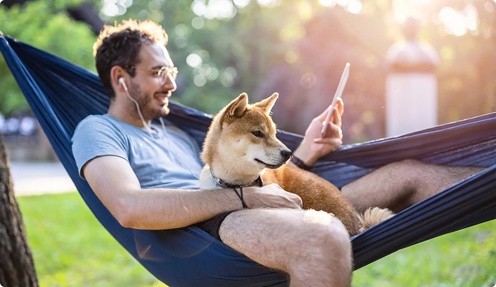 Muž oddychuje v hojdacej sieti v parku, počúva hudbu cez slúchadlá a používa tablet, zatiaľ čo vedľa neho sedí pes.