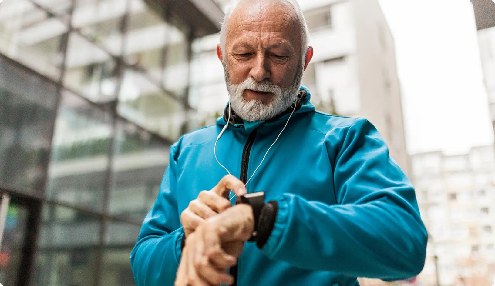 Starší muž v športovej bunde stojí vonku v meste a pozerá sa na smart hodinky na svojom zápästí, pričom má v ušiach slúchadlá.