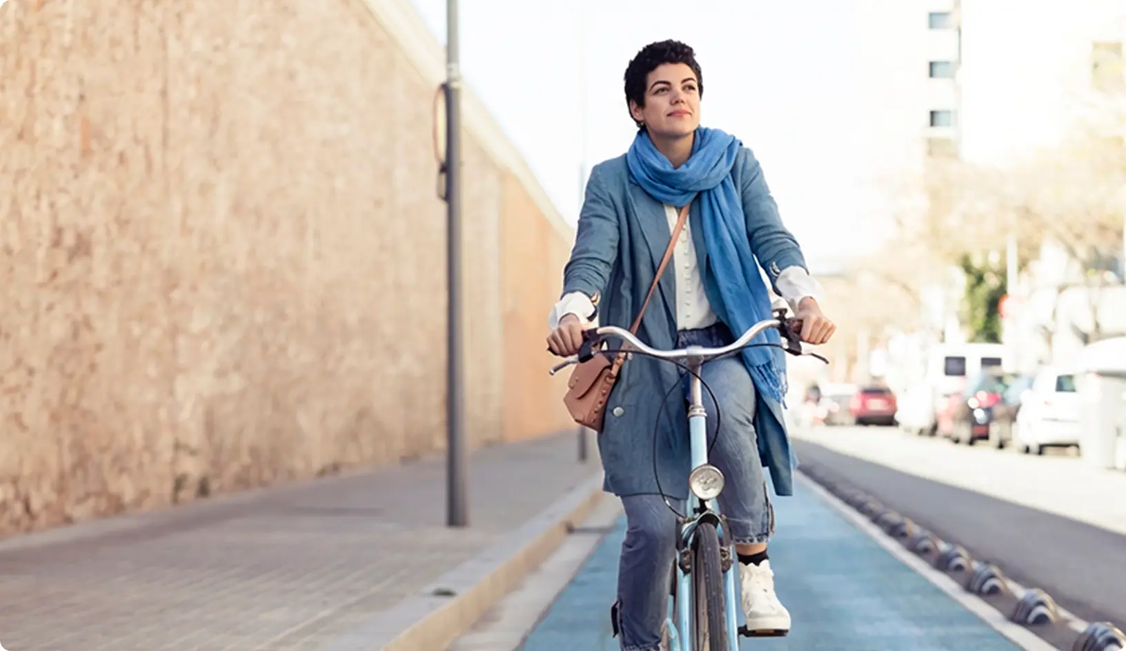 Mladá žena jazdí na bicykli po mestskom cyklochodníku, oblečená v kabáte a modrom šále, s kabelkou prevesenou cez plece.