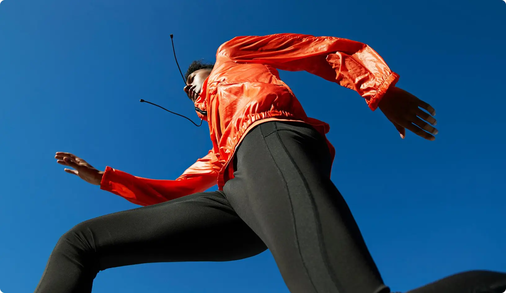 Osoba v oranžovej športovej bunde a čiernych legínach beží vonku, snímaná z nízkeho uhla proti modrej oblohe.