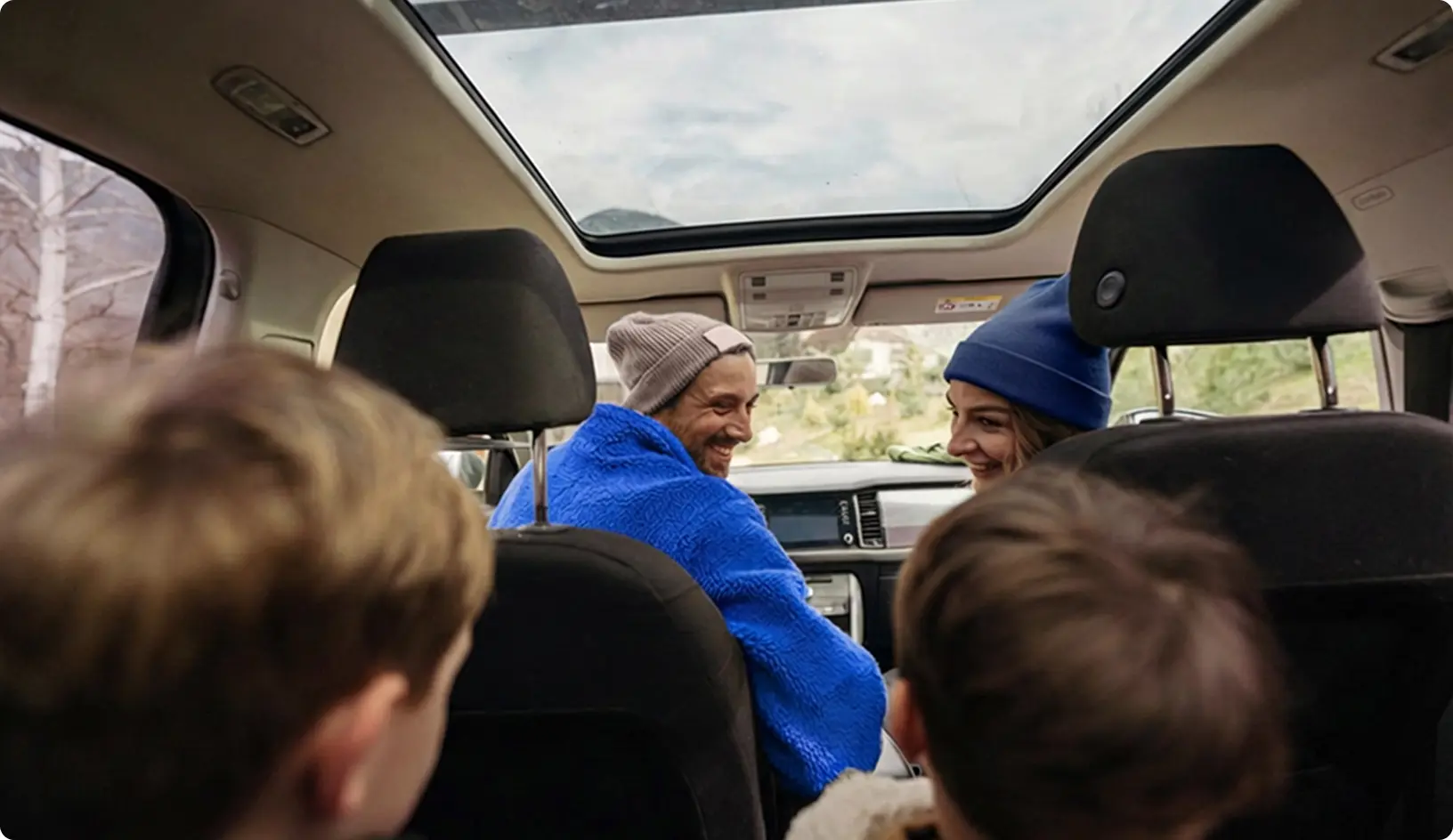 Rodina sedí v aute počas jazdy, rodičia sa s úsmevom otáčajú dozadu k deťom na zadných sedadlách.